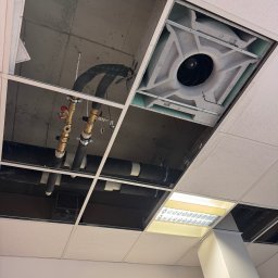 Otwarty sufit podwieszany z widocznymi rurami, zaworami i elementami wentylacji. Widoczne instalacje nad sufitem, oświetlenie i fragment ściany z zegarem.