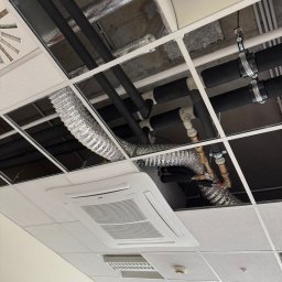 Instalacja rur i przewodów wentylacyjnych podwieszonych pod sufitem rastrowym w biurze. Widoczny klimatyzator kasetonowy i elementy instalacji.