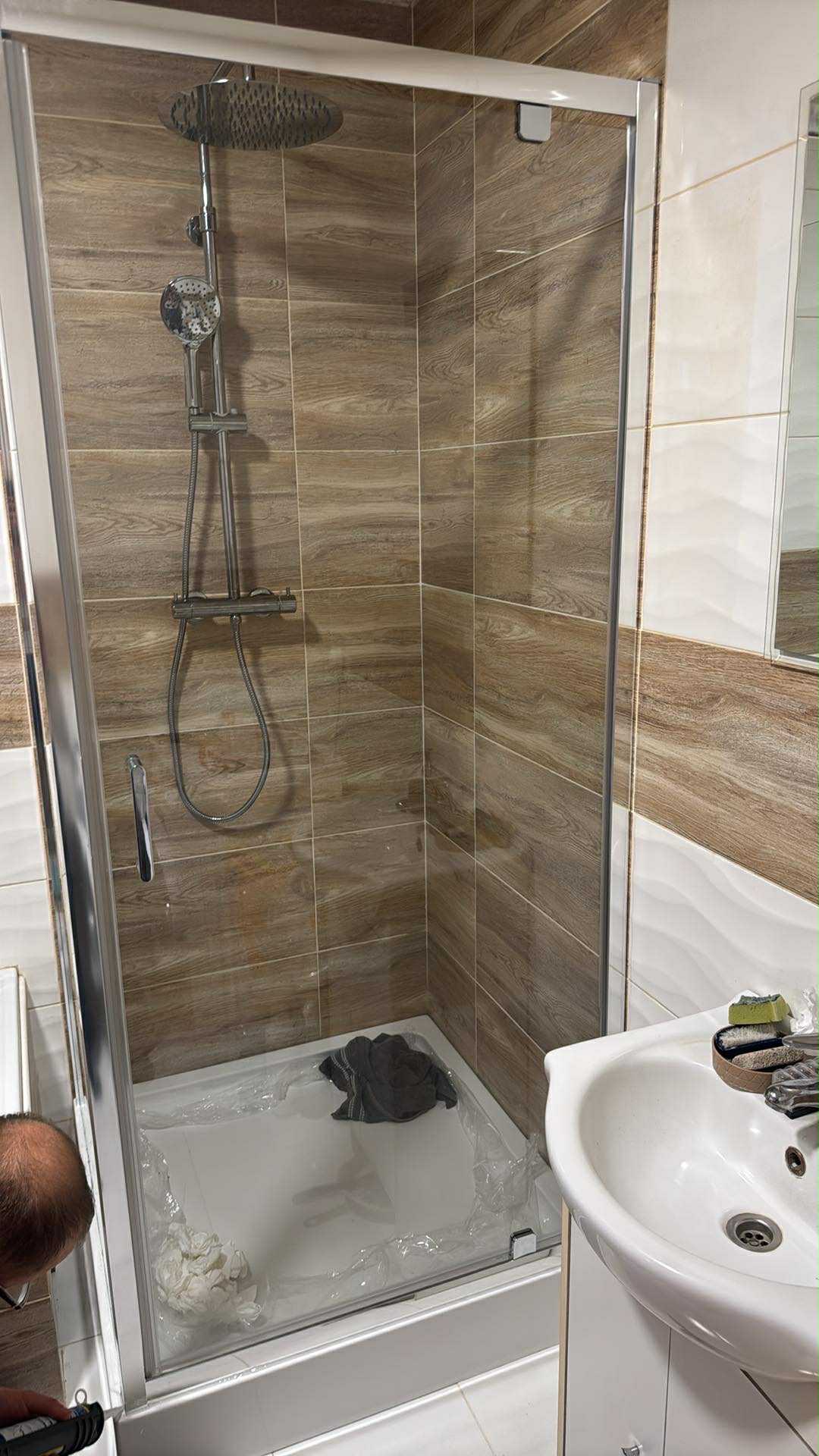 Nowa kabina prysznicowa z deszczownicą, zabezpieczona folią ochronną, w łazience z brązowo-białymi płytkami imitującymi drewno. Widoczna umywalka i fragment osoby.