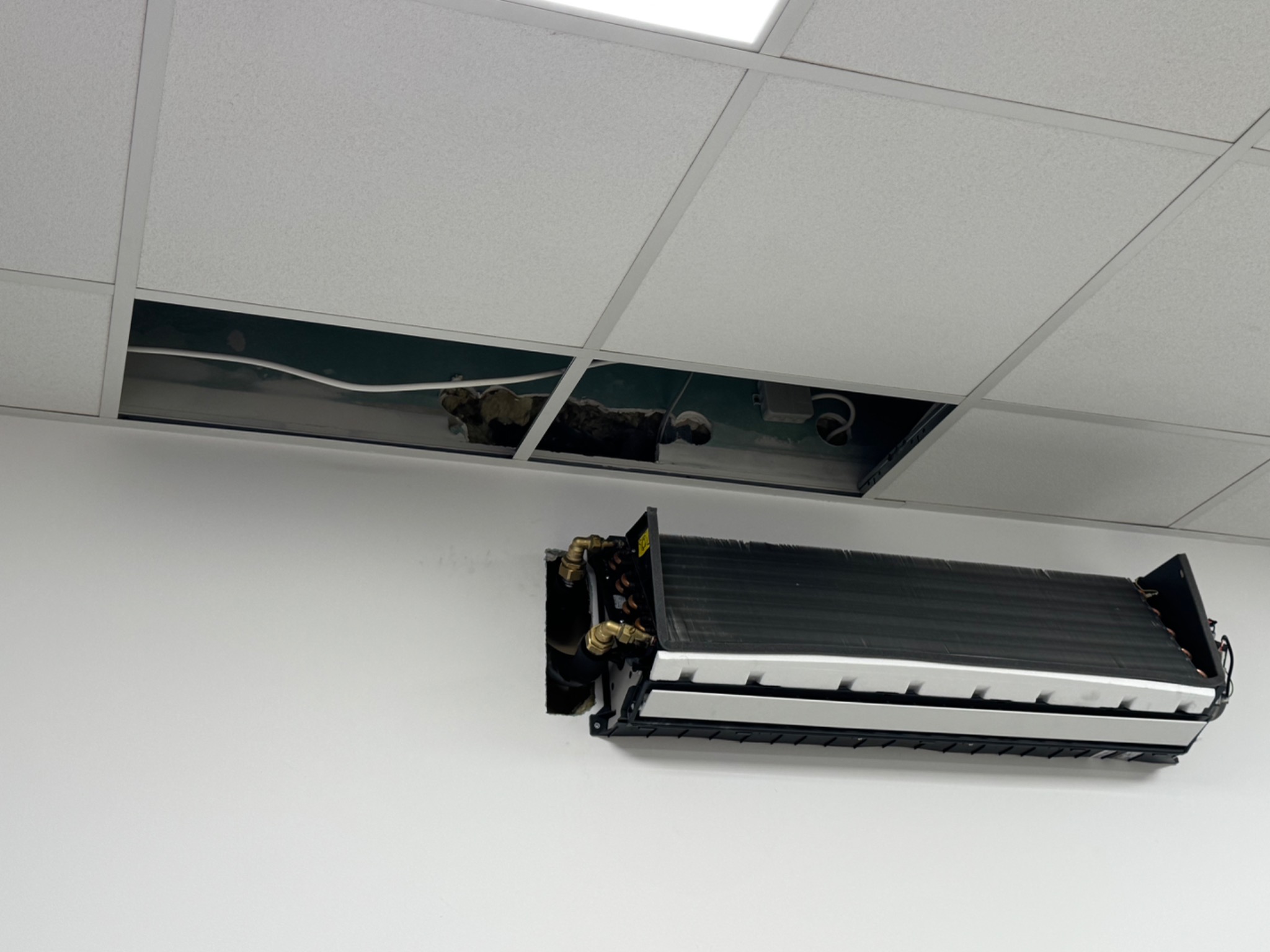 Klimatyzator ścienny z widocznymi miedzianymi rurami, zamontowany na białej ścianie. W tle sufit podwieszany z uszkodzonym panelem.