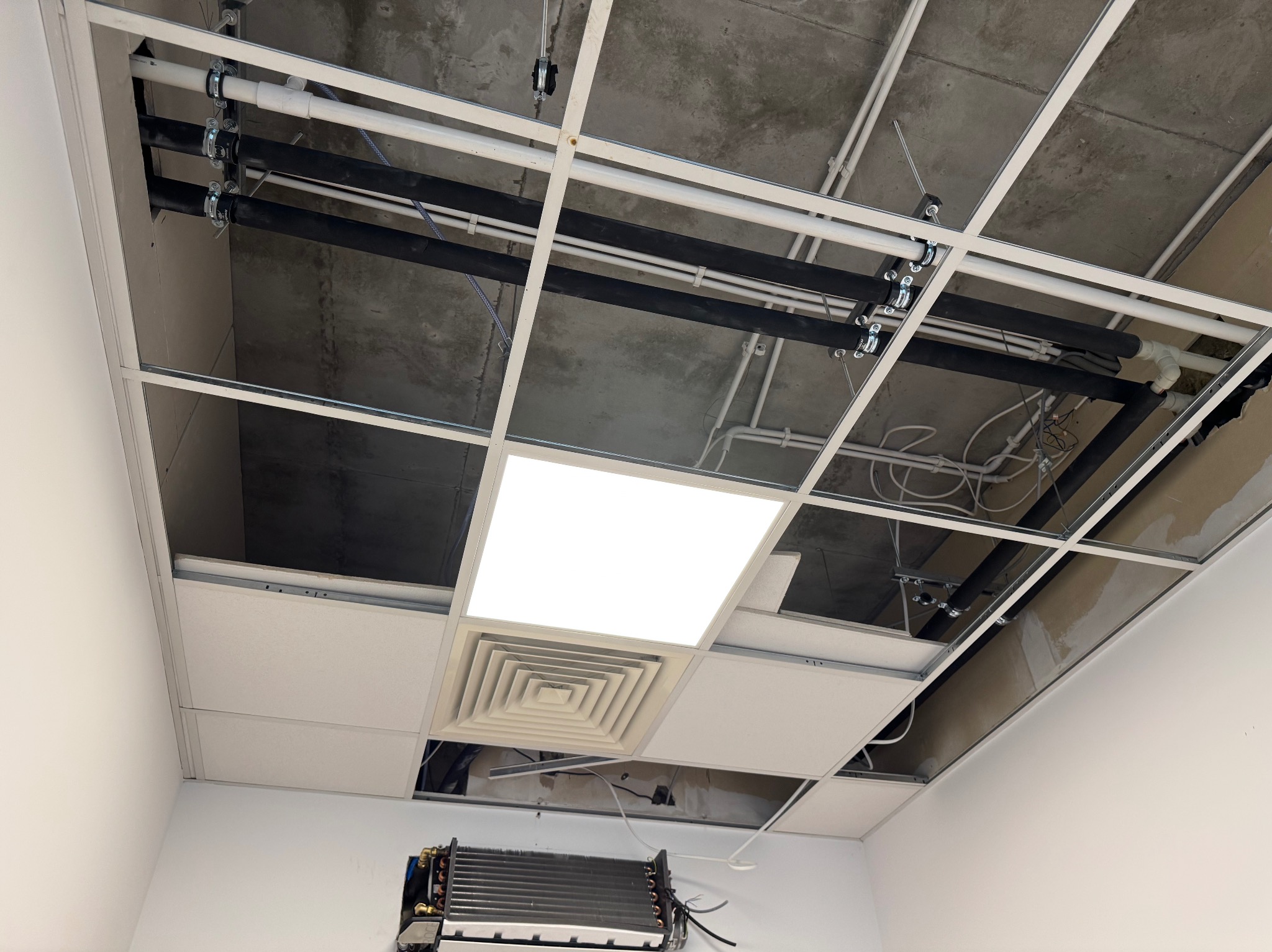 Instalacja rur podwieszonych pod sufitem rastrowym z widoczną klimatyzacją i oświetleniem. Widok z dołu, ukazujący elementy instalacyjne w przestrzeni międzystropowej.