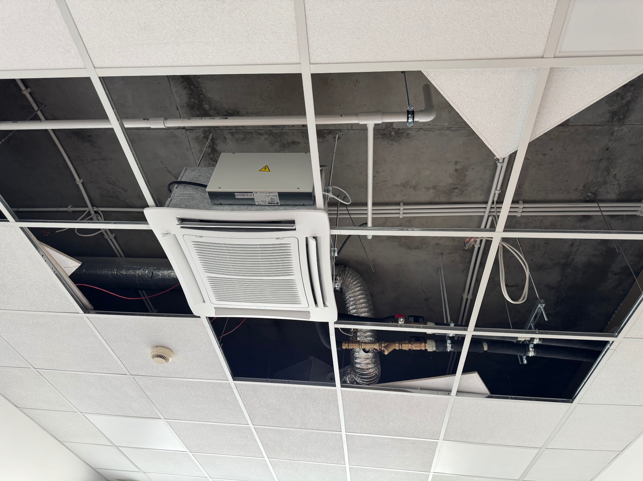 Kasetonowy sufit podwieszany z widoczną instalacją klimatyzacji, rurami i przewodami. Część kasetonów jest zdjęta, odsłaniając elementy instalacyjne.
