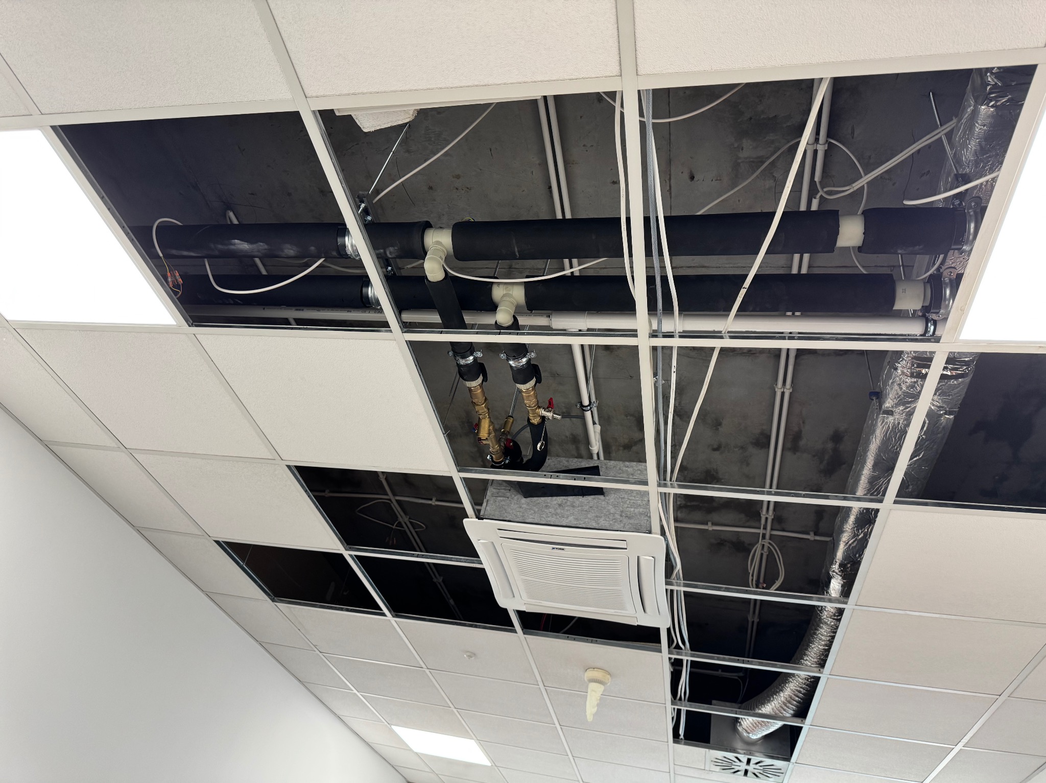 Instalacja rur i klimatyzacji w przestrzeni sufitu podwieszanego. Widoczne rury w izolacji, elementy instalacji hydraulicznej i klimatyzator kasetonowy.