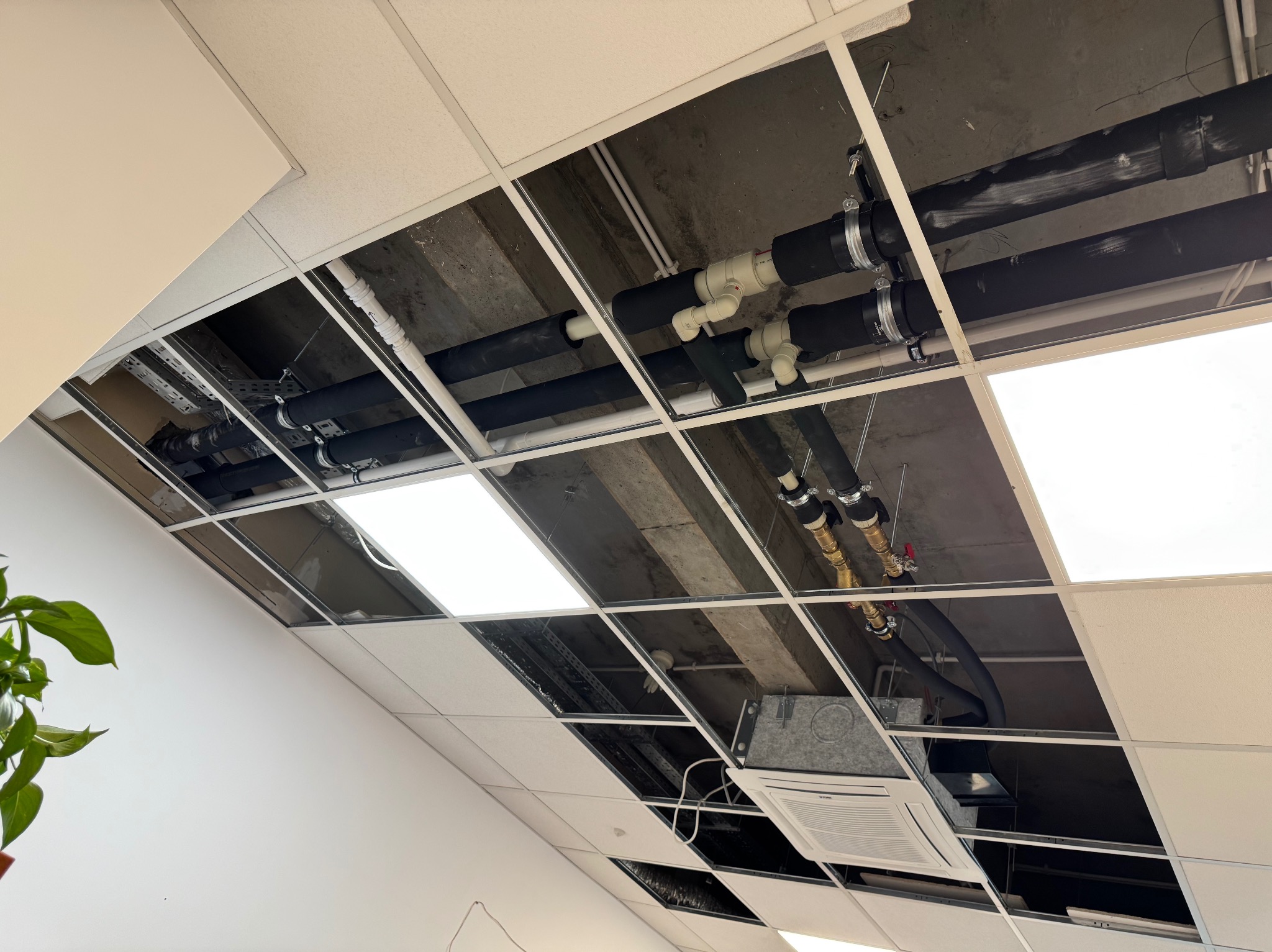 Instalacja rur i klimatyzacji pod sufitem podwieszanym w biurze. Widoczne czarne, białe i miedziane rury z zaworami. Część paneli sufitowych jest zdjęta.