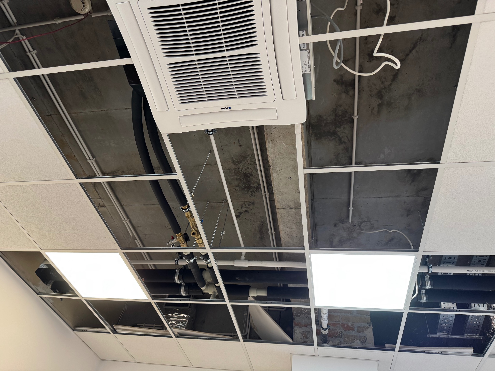 Kasetonowy sufit podwieszany z widoczną instalacją klimatyzacji i rurami. Widoczne elementy instalacji nad sufitem, w tym rury w izolacji i biała jednostka klimatyzacyjna.