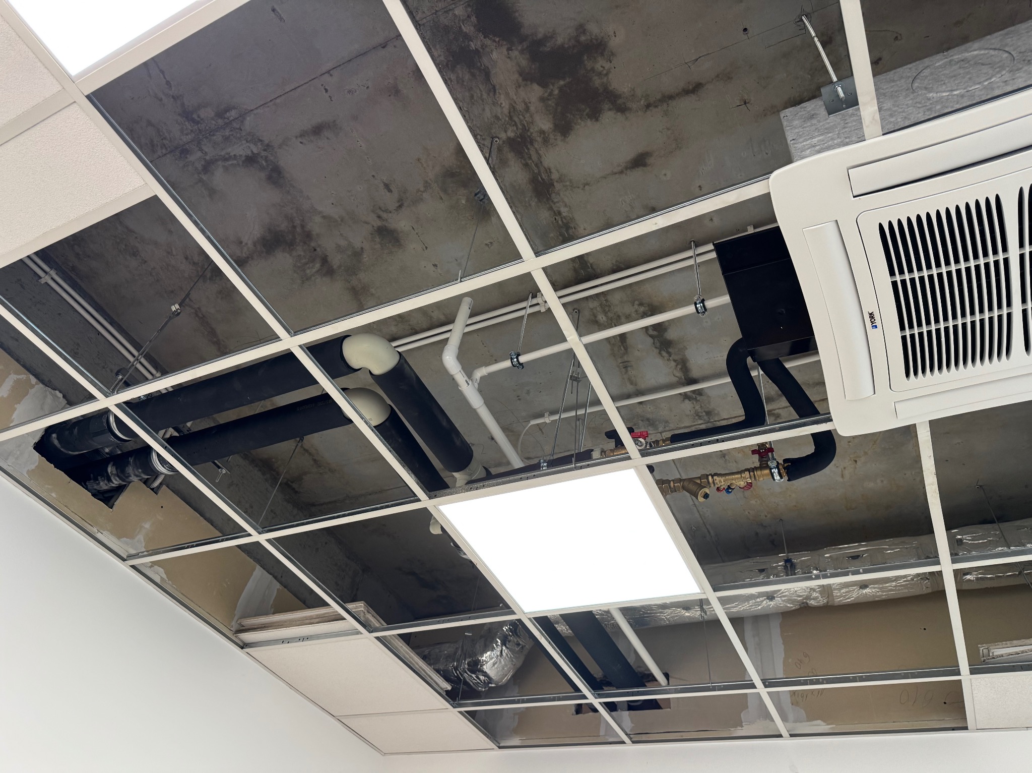Widok na sufit podwieszany z widocznymi instalacjami: rury, klimatyzacja York, oświetlenie LED. Elementy instalacji hydraulicznej i wentylacyjnej.