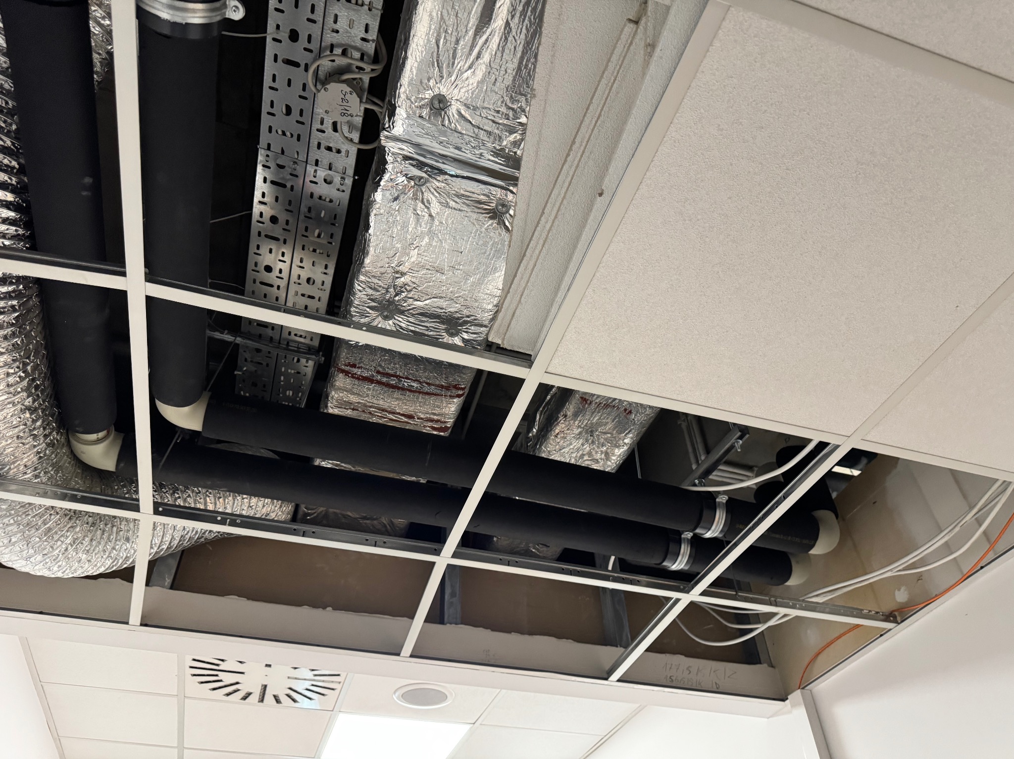 Instalacja wentylacyjna i rury w przestrzeni podwieszanego sufitu biurowego, widoczne elementy mocujące i izolacja termiczna.