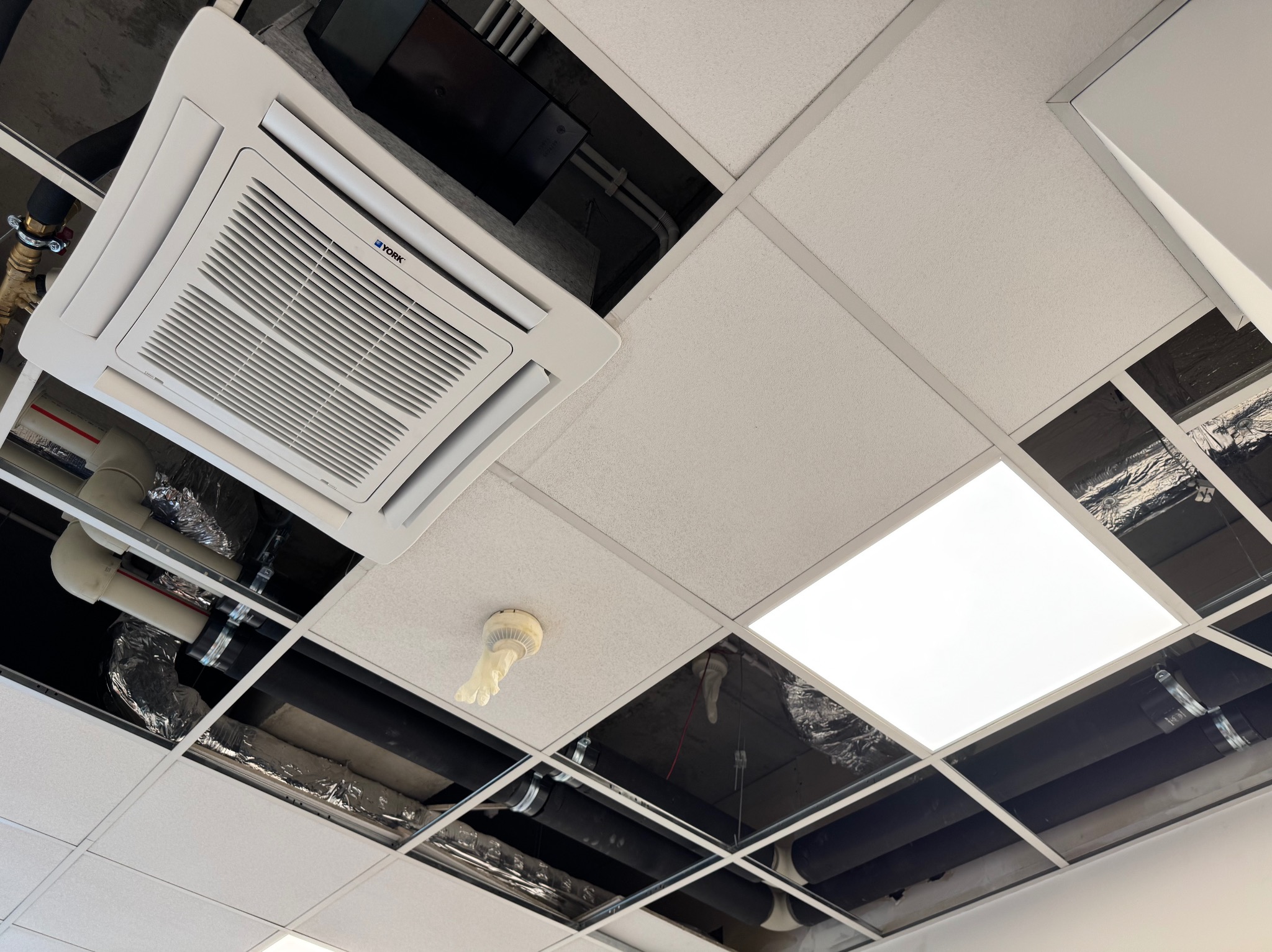 Kasetonowy sufit z widoczną instalacją klimatyzacji York, rurami, oświetleniem LED i czujnikiem dymu. Widok z dołu, instalacja w trakcie montażu.