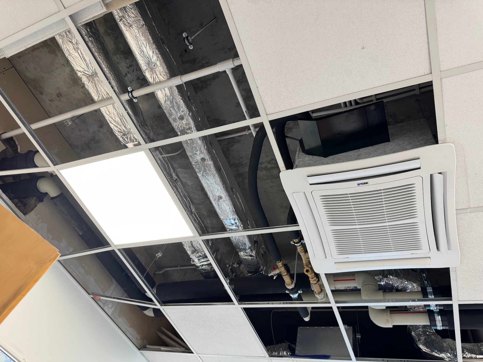 Widok instalacji podwieszanej klimatyzacji YORK z widocznymi rurami i przewodami wentylacyjnymi, zamontowanej w przestrzeni sufitu modułowego.