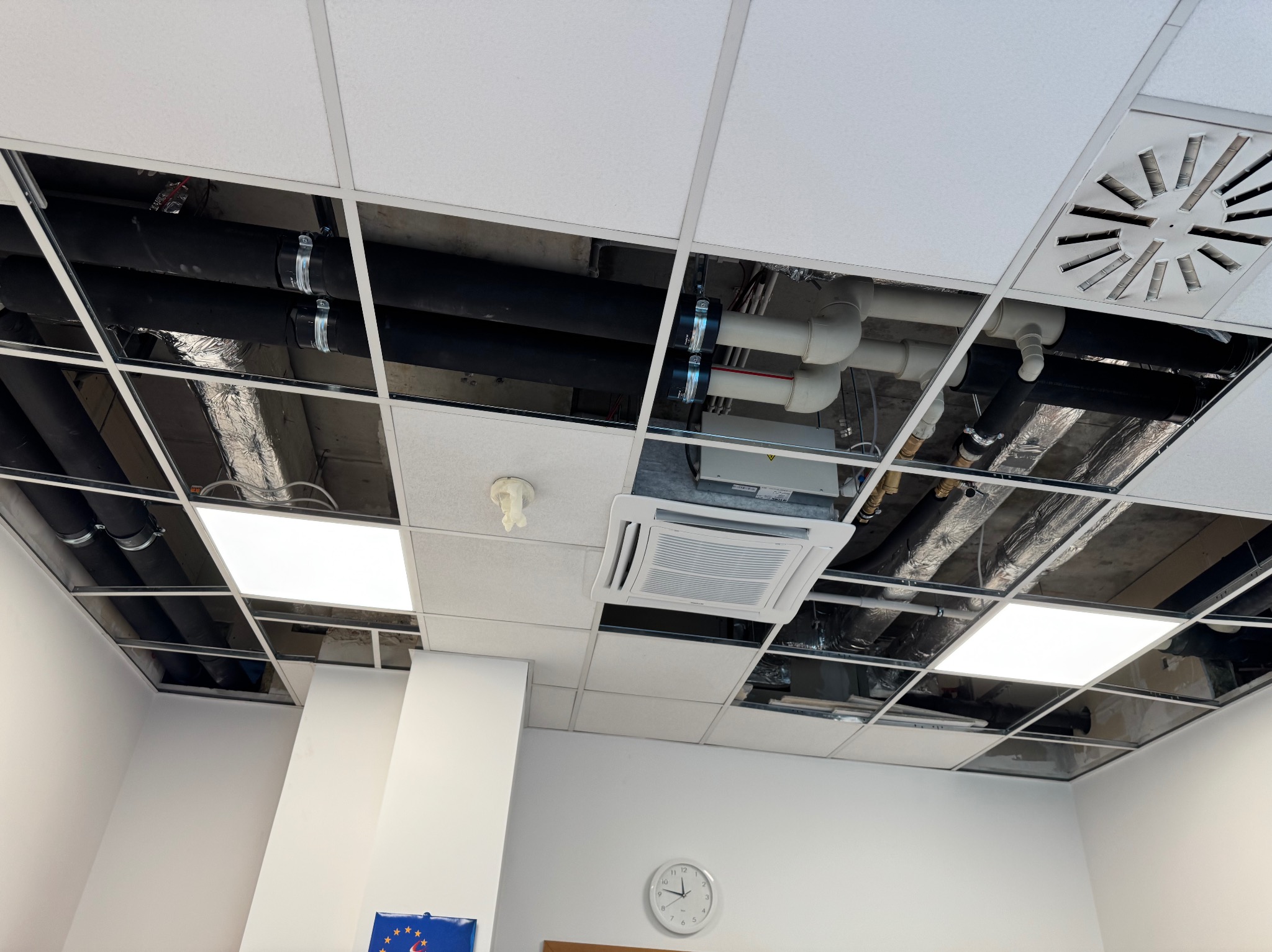 Widok na sufit podwieszany z widocznymi instalacjami: rury, wentylacja, klimatyzacja i oświetlenie. Układ instalacji w przestrzeni nad sufitem.