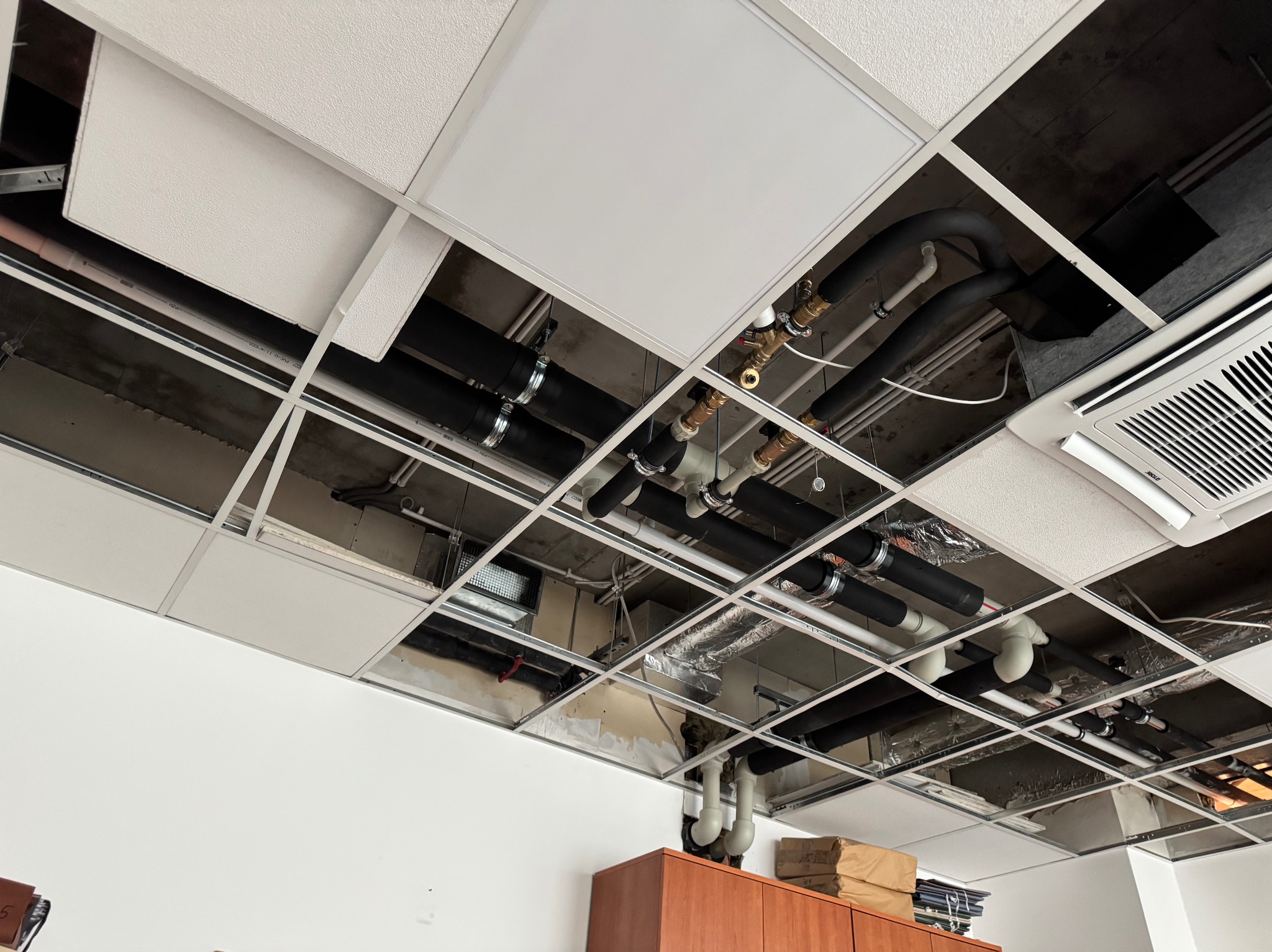 Instalacje sanitarne i wentylacyjne pod sufitem podwieszanym z widocznymi rurami, przewodami i klimatyzacją. Widok z dołu na instalacje.