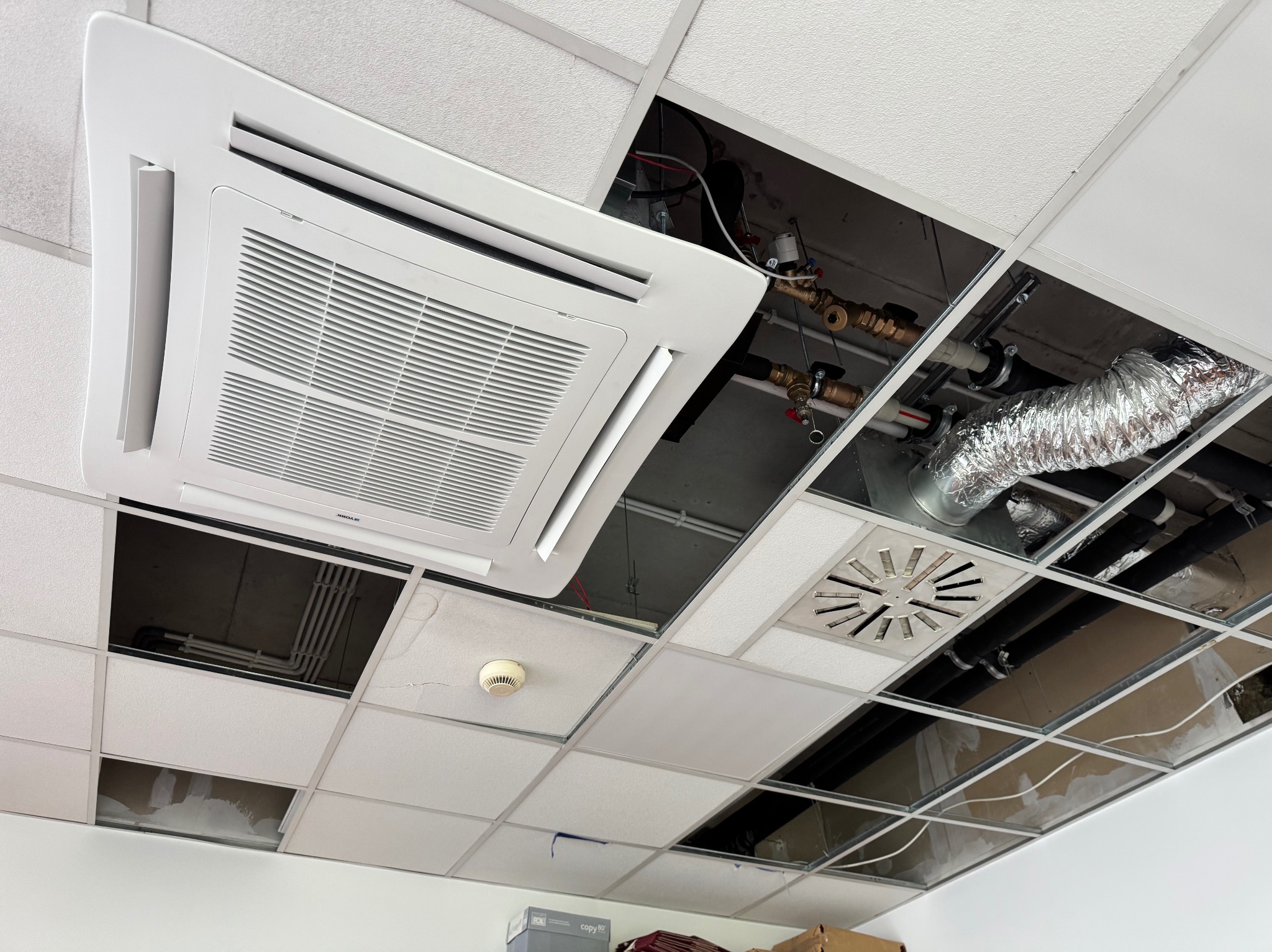 Kasetonowy sufit z widoczną instalacją klimatyzacji i wentylacji. Część kasetonów zdjęta, ukazując rury i przewody. Widoczny klimatyzator kasetonowy.