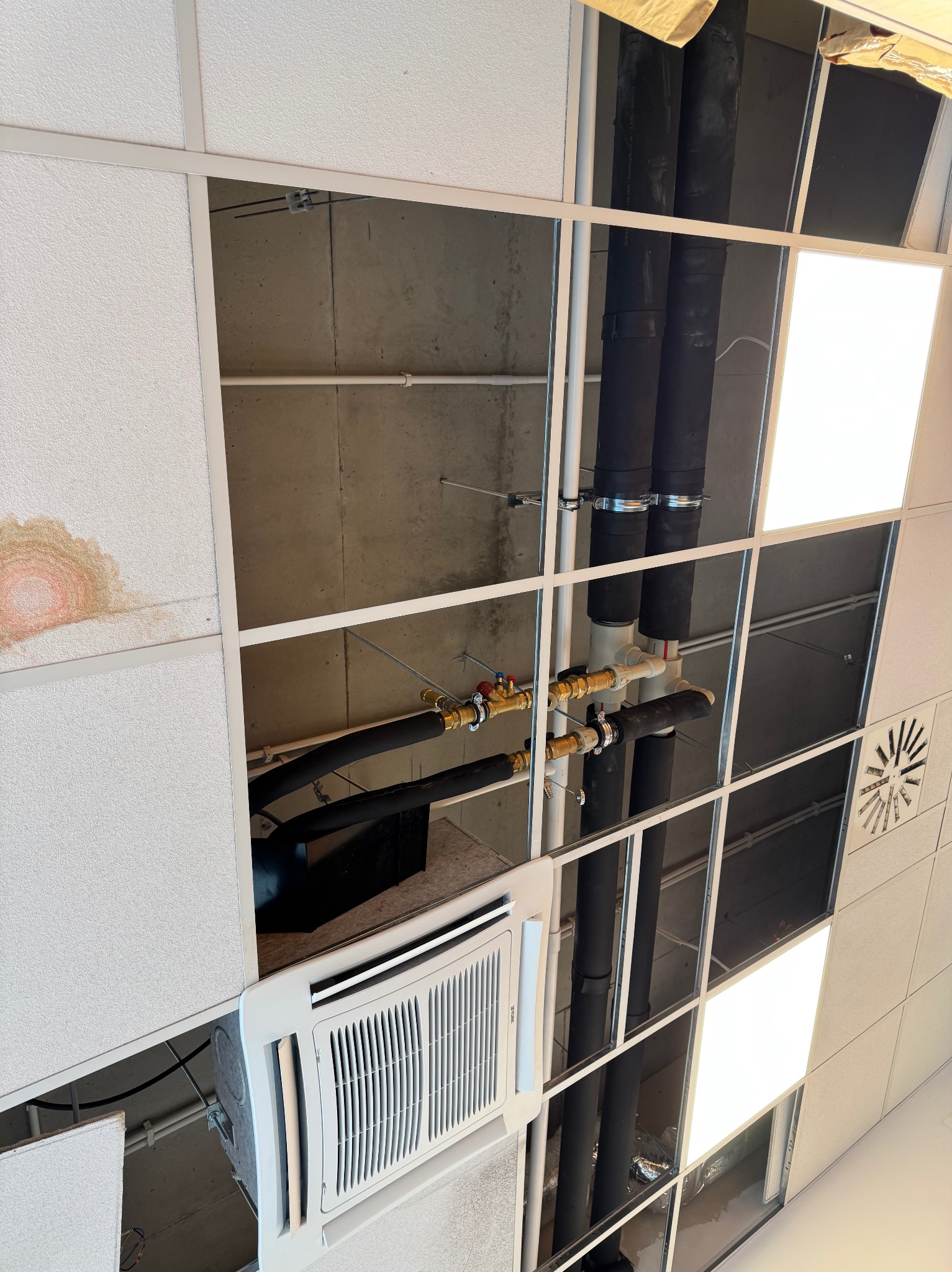 Widok instalacji podwieszonych pod sufitem rastrowym: rury, klimatyzacja kasetonowa, oświetlenie. Widoczne elementy instalacji hydraulicznej i wentylacyjnej.