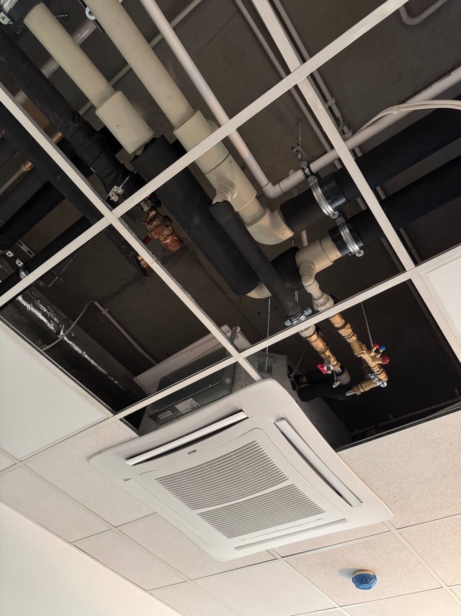Instalacja rur i zaworów pod sufitem podwieszanym, widoczny klimatyzator kasetonowy. Różne materiały rur: czarne izolowane, białe PCV, miedziane z zaworami.