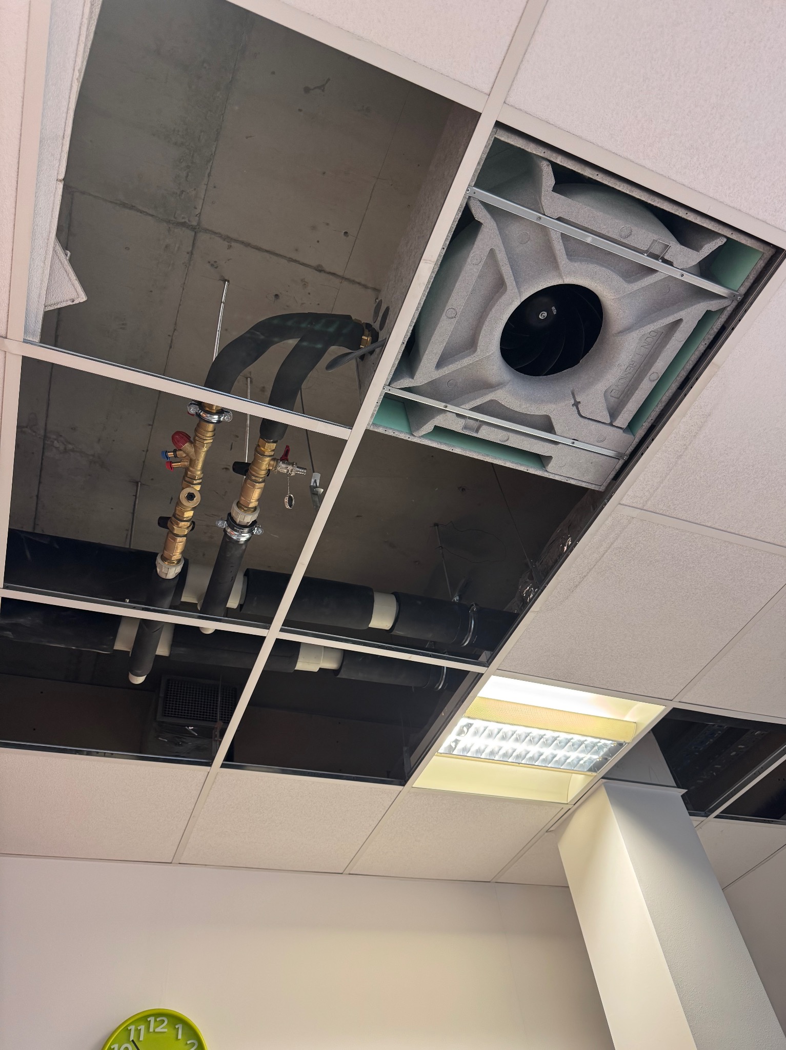 Otwarty sufit podwieszany z widocznymi rurami, zaworami i elementami wentylacji. Widoczne instalacje nad sufitem, oświetlenie i fragment ściany z zegarem.