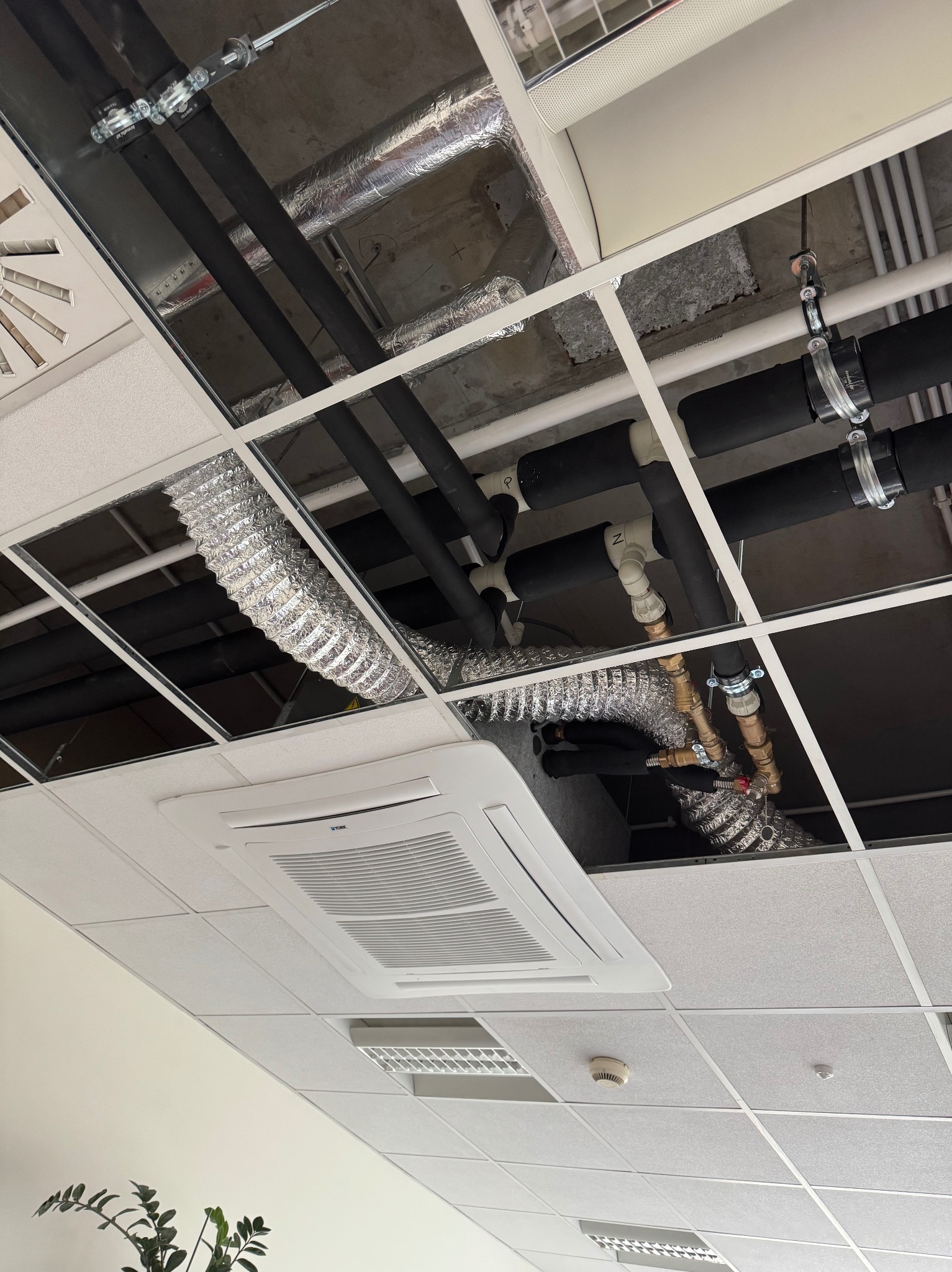 Instalacja rur i przewodów wentylacyjnych podwieszonych pod sufitem rastrowym w biurze. Widoczny klimatyzator kasetonowy i elementy instalacji.