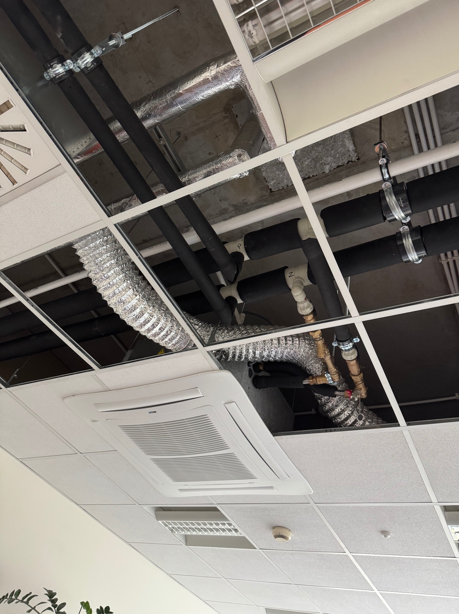 Widok na instalację wentylacyjną i rury pod sufitem podwieszanym w biurze, z widocznym klimatyzatorem kasetonowym i elementami mocującymi.
