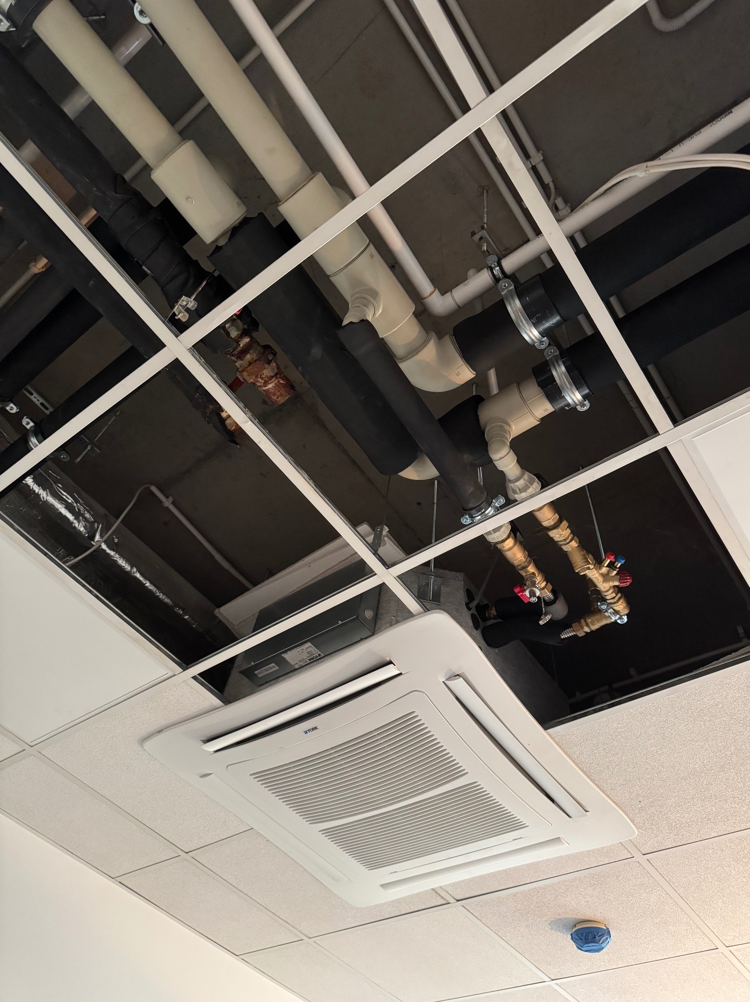 Instalacje sanitarne i klimatyzacja pod sufitem podwieszanym: rury, zawory, klimatyzator kasetonowy. Widok z dołu, oświetlenie biurowe.