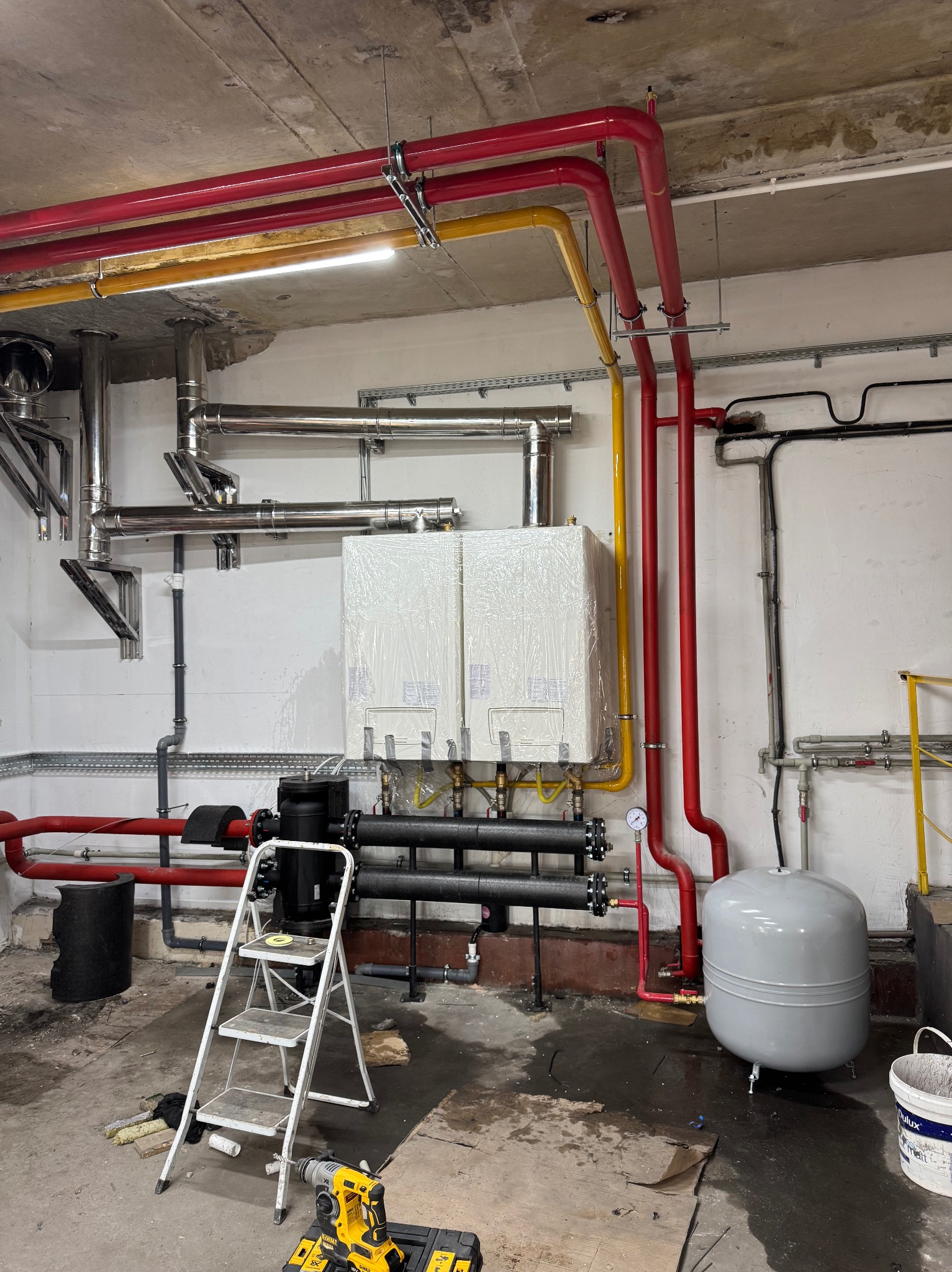 W kotłowni dwa białe piece CO w folii, z podłączonymi rurami (czerwone, żółte, czarne, srebrne). Na podłodze drabina i narzędzia. Widoczne elementy instalacji.