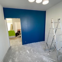 FORTISANIMA MARCIN ANTCZAK - Świeżo pomalowana na niebiesko ściana z widocznymi śladami farby na podłodze. Obok stoi aluminiowa drabina, a nad ścianą wiszą trzy białe lampy.