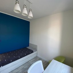 FORTISANIMA MARCIN ANTCZAK - Pokój po malowaniu: granatowa ściana za łóżkiem z wzorzystą pościelą, białe ściany, lampa sufitowa z trzema kloszami, jasne panele podłogowe i białe meble.