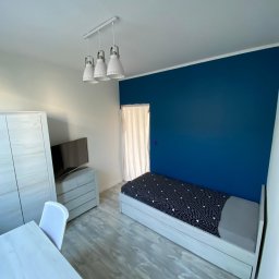 FORTISANIMA MARCIN ANTCZAK - Pokój po malowaniu: intensywnie niebieska ściana za łóżkiem z wzorzystą pościelą. Białe meble, podłoga imitująca drewno i nowoczesna lampa wisząca.