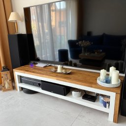 FORTISANIMA MARCIN ANTCZAK - Drewniany stolik RTV z białymi nogami, na którym stoi duży telewizor. Na blacie dekoracje, a pod spodem sprzęt audio. Widoczne kolumny głośnikowe i zasłony w tle.