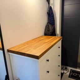 FORTISANIMA MARCIN ANTCZAK - Biała komoda z drewnianym blatem w przedpokoju, obok czarne drzwi i wieszak na ubrania. Widoczne buty i wycieraczka z napisem 'Hello!'.