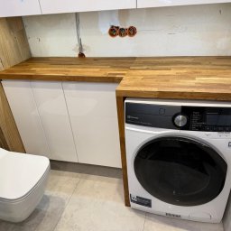 FORTISANIMA MARCIN ANTCZAK - Drewniany blat nad białą szafką i pralką Electrolux w łazience z beżowymi płytkami. Widoczne gniazdka elektryczne i wisząca toaleta. Nowoczesne wykończenie.