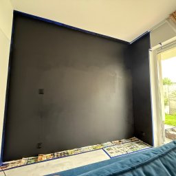 FORTISANIMA MARCIN ANTCZAK - Ściana w trakcie malowania na ciemny kolor, zabezpieczona taśmą malarską i gazetami. Widoczna drabina malarska i fragment sofy. Prace malarskie w pomieszczeniu z oknem.