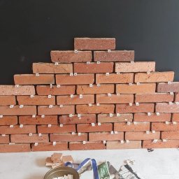 FORTISANIMA MARCIN ANTCZAK - Układanie dekoracyjnej ściany z cegieł w kształcie piramidy na ciemnym tle. Widoczne plastikowe krzyżyki dystansowe i wiadro z zaprawą. Prace wykończeniowe w trakcie.