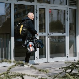 Marcin Antczak Naprawy domowe - Mężczyzna z plecakiem i torbą narzędziową Bosch, idący przed przeszklonym wejściem do budynku. Widoczny odkurzacz przemysłowy. Usługi remontowe, instalacyjne.
