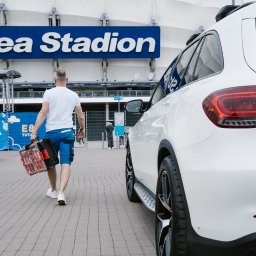 Marcin Antczak Naprawy domowe - Mężczyzna z skrzynką narzędzi idzie w stronę stadionu Enea, obok zaparkowany biały samochód. Widoczny fragment czarnego auta po lewej stronie.