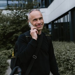 Marcin Antczak Naprawy domowe - Uśmiechnięty mężczyzna z odkurzaczem na plecach rozmawia przez telefon na tle nowoczesnego budynku biurowego i zieleni.