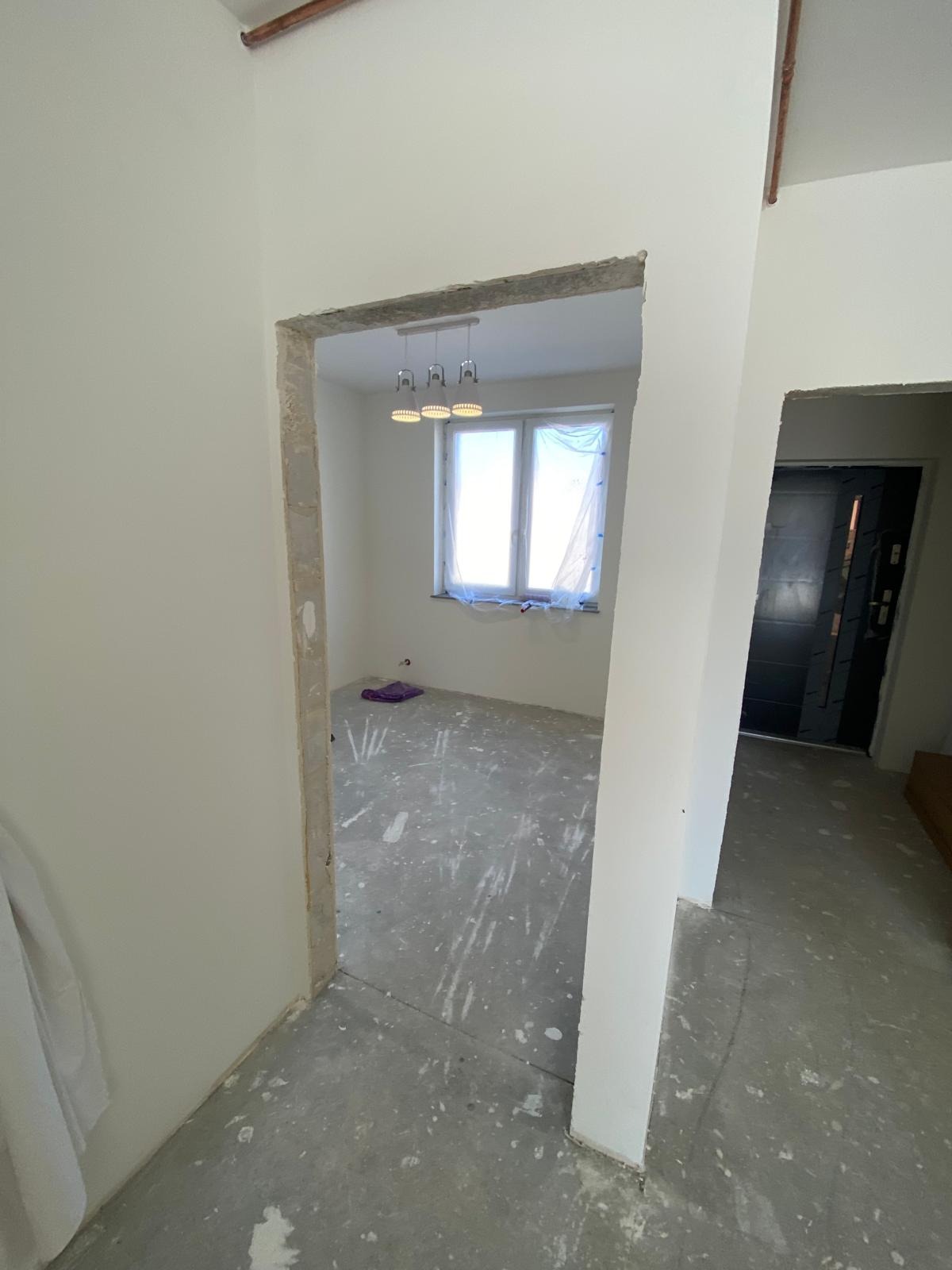 Wnętrze w trakcie remontu: białe ściany, surowy beton na podłodze z plamami farby, okno zabezpieczone folią i wisząca lampa. Widok z perspektywy otwartych drzwi.