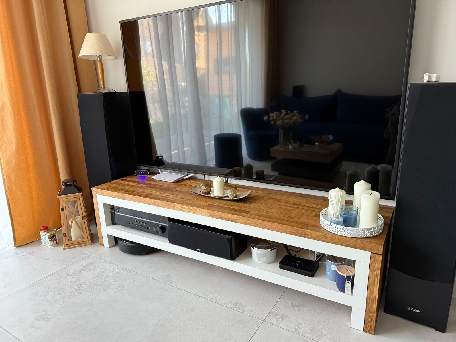 Drewniany stolik RTV z białymi nogami, na którym stoi duży telewizor. Na blacie dekoracje, a pod spodem sprzęt audio. Widoczne kolumny głośnikowe i zasłony w tle.