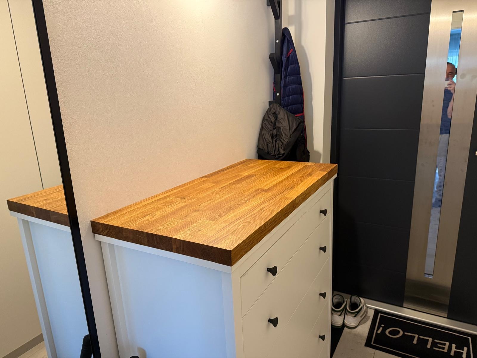 Biała komoda z drewnianym blatem w przedpokoju, obok czarne drzwi i wieszak na ubrania. Widoczne buty i wycieraczka z napisem 'Hello!'.