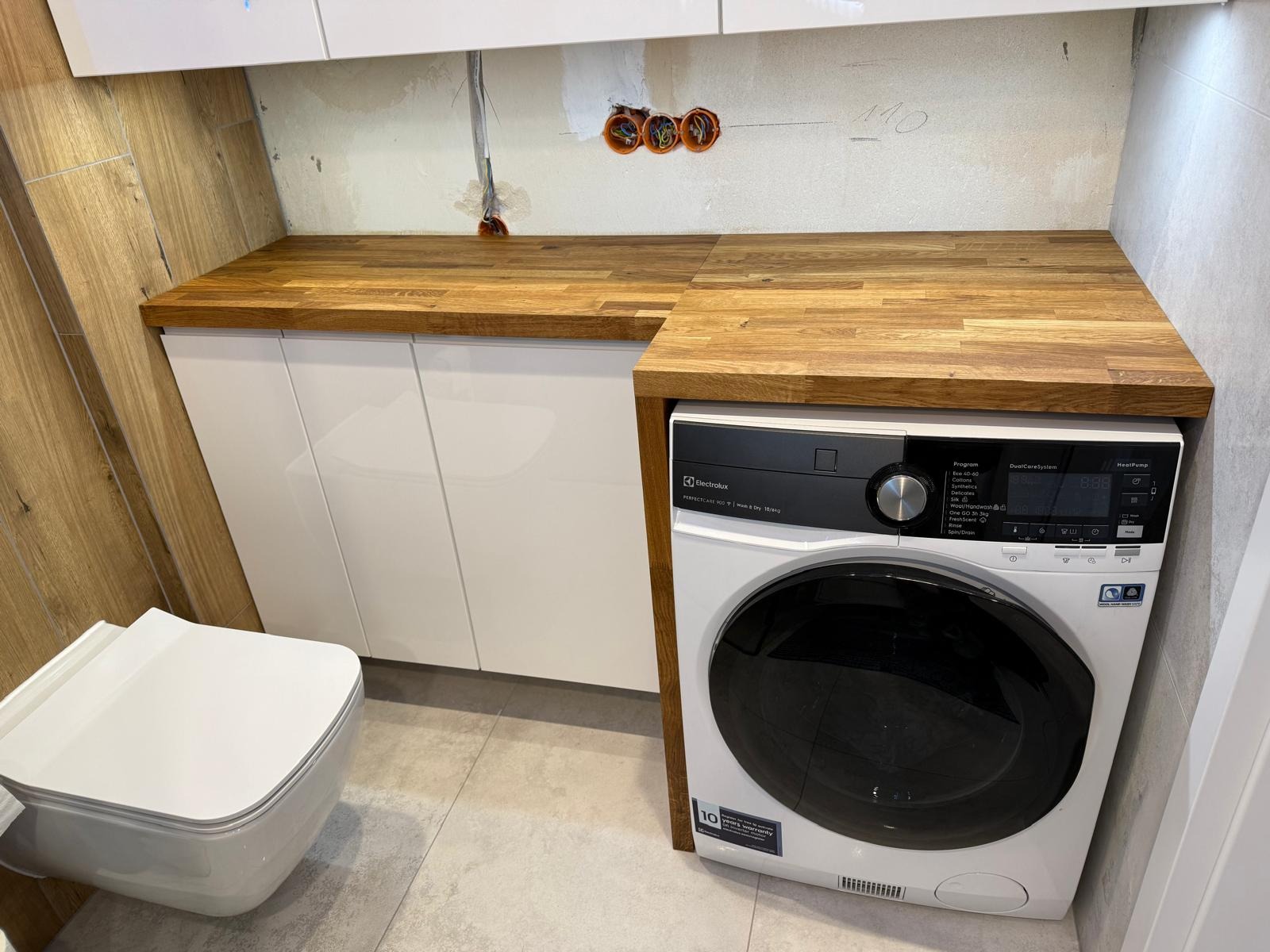Drewniany blat nad białą szafką i pralką Electrolux w łazience z beżowymi płytkami. Widoczne gniazdka elektryczne i wisząca toaleta. Nowoczesne wykończenie.