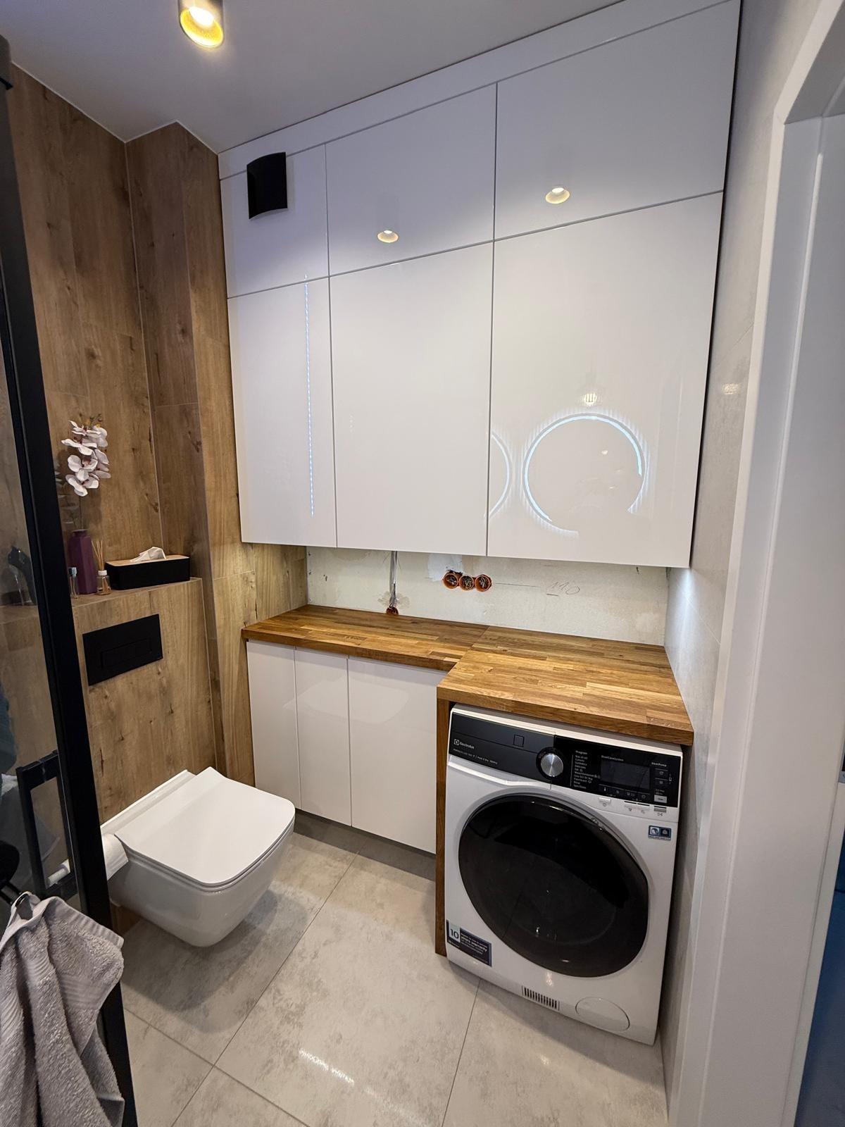 Nowoczesna łazienka z drewnianym blatem, białą zabudową meblową i pralką Electrolux. Widoczne gniazdka elektryczne nad blatem. Minimalistyczny design.