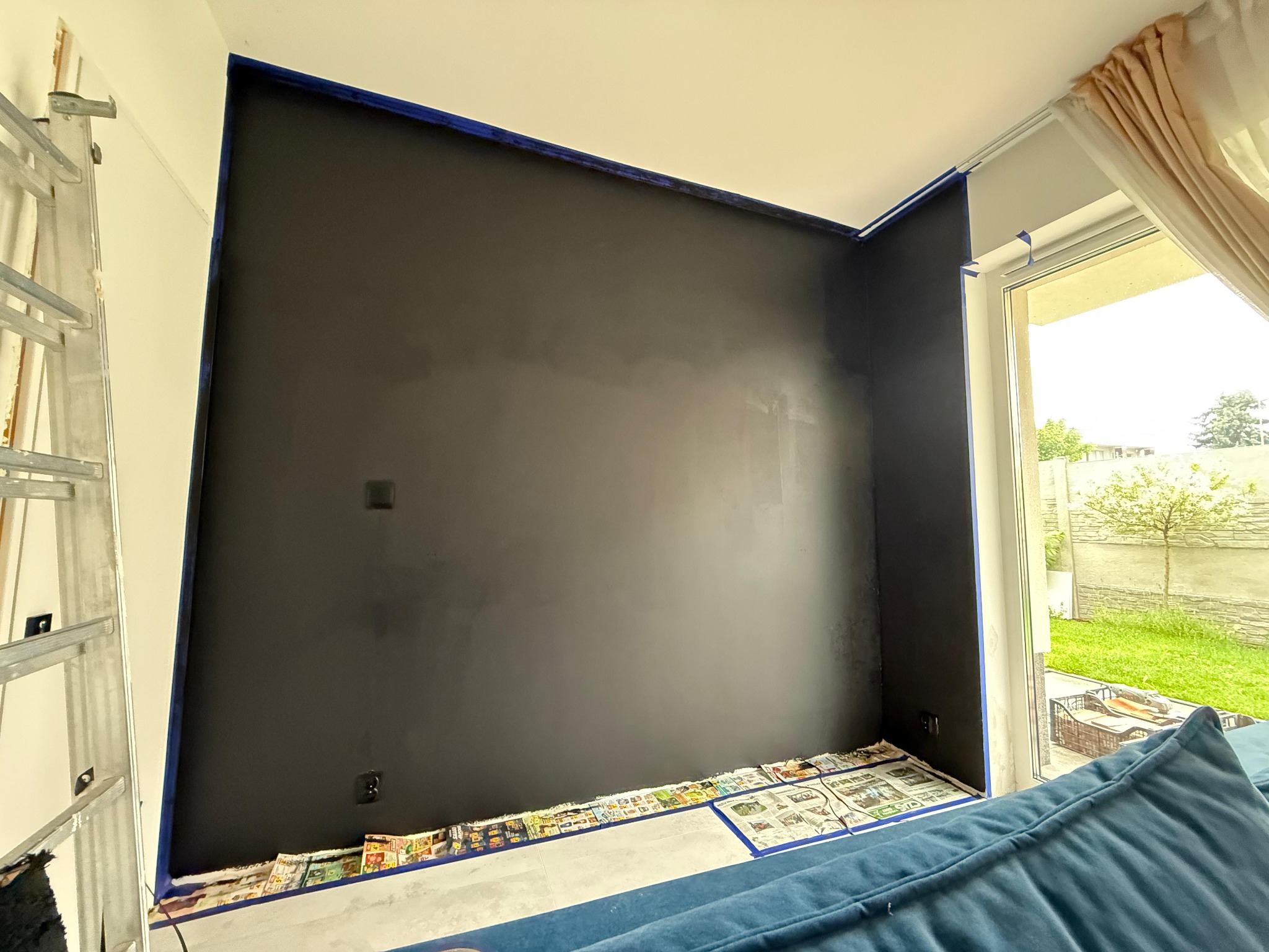 Ściana w trakcie malowania na ciemny kolor, zabezpieczona taśmą malarską i gazetami. Widoczna drabina malarska i fragment sofy. Prace malarskie w pomieszczeniu z oknem.