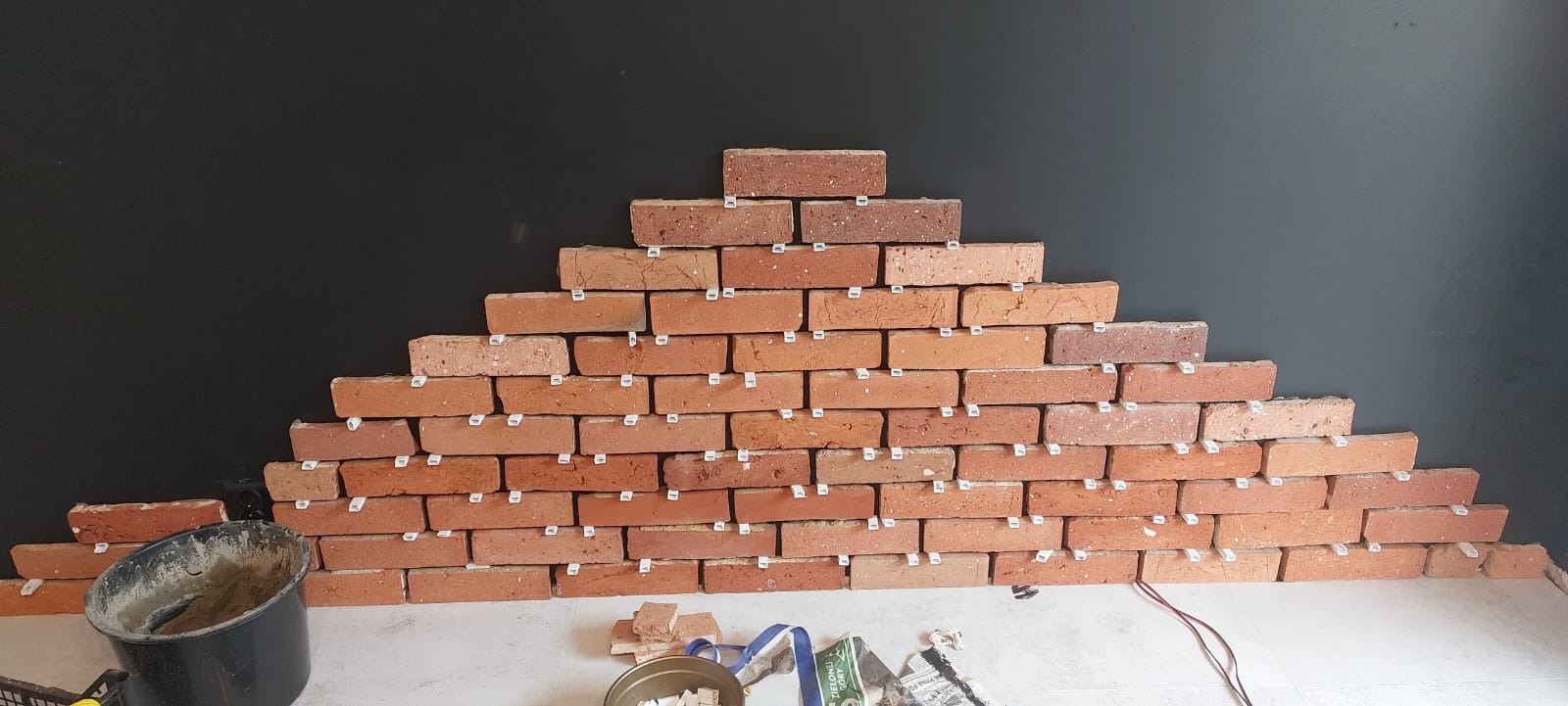 Układanie dekoracyjnej ściany z cegieł w kształcie piramidy na ciemnym tle. Widoczne plastikowe krzyżyki dystansowe i wiadro z zaprawą. Prace wykończeniowe w trakcie.