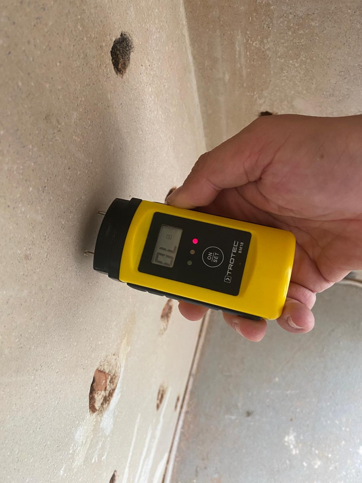 Pomiar wilgotności ściany za pomocą wilgotnościomierza Trotec BM18. Widoczne otwory w ścianie. Urządzenie w dłoni, wyświetlacz pokazuje wartość.