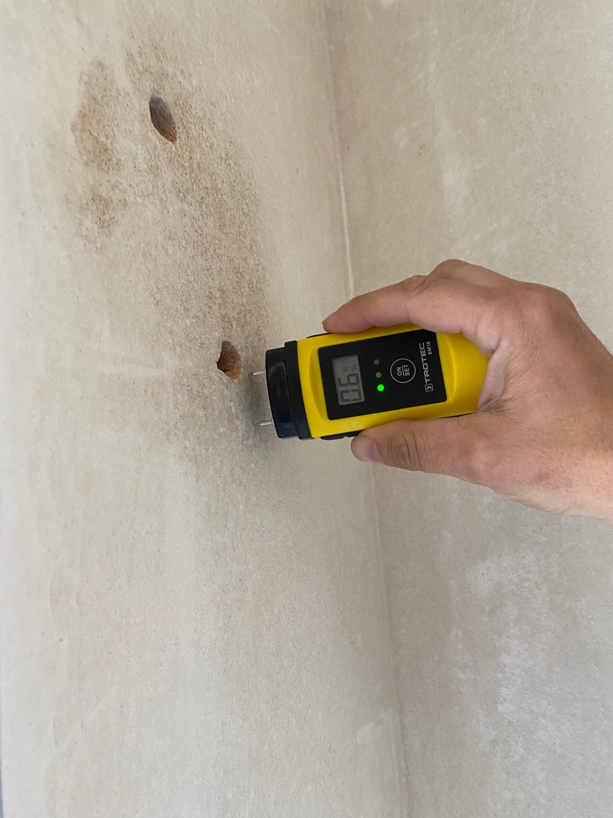 Pomiar wilgotności ściany z widocznymi plamami i otworami, przy użyciu żółtego wilgotnościomierza. Urządzenie w dłoni, wyświetla wynik pomiaru.