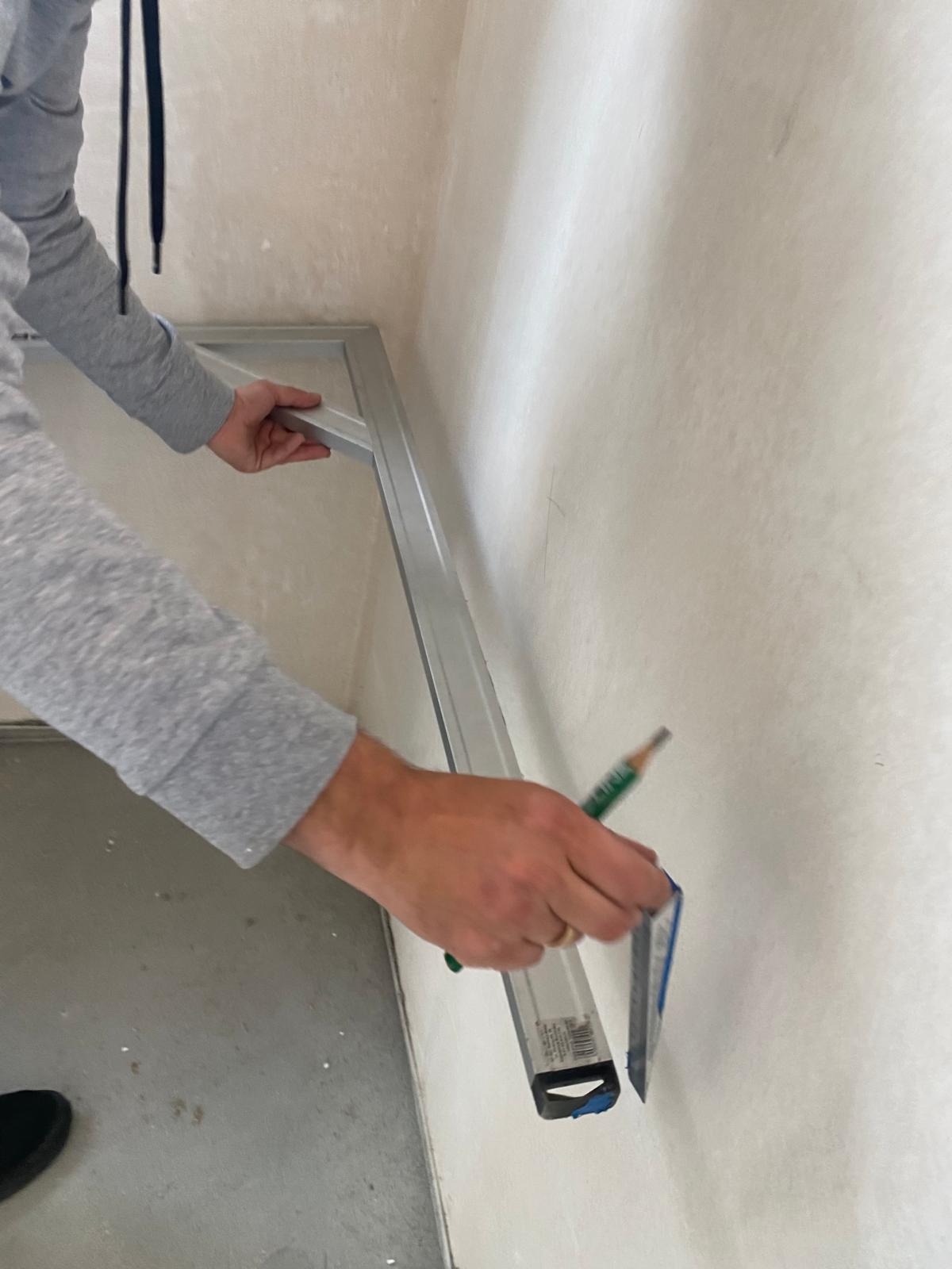 Pracownik przykłada poziomicę do ściany i zaznacza ołówkiem punkt, przygotowując się do montażu. Widoczny fragment metalowej konstrukcji i surowa ściana.