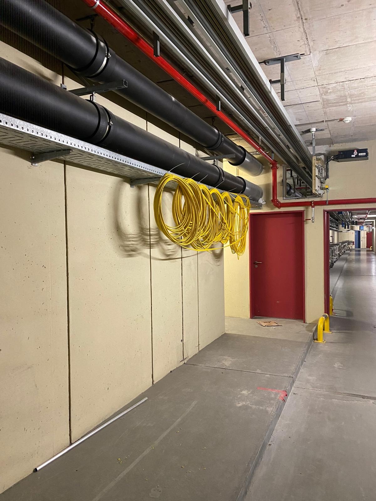 Żółte przewody zwisają z metalowej półki pod sufitem, obok rur i czerwonych drzwi w korytarzu. Widok na instalacje w budynku.