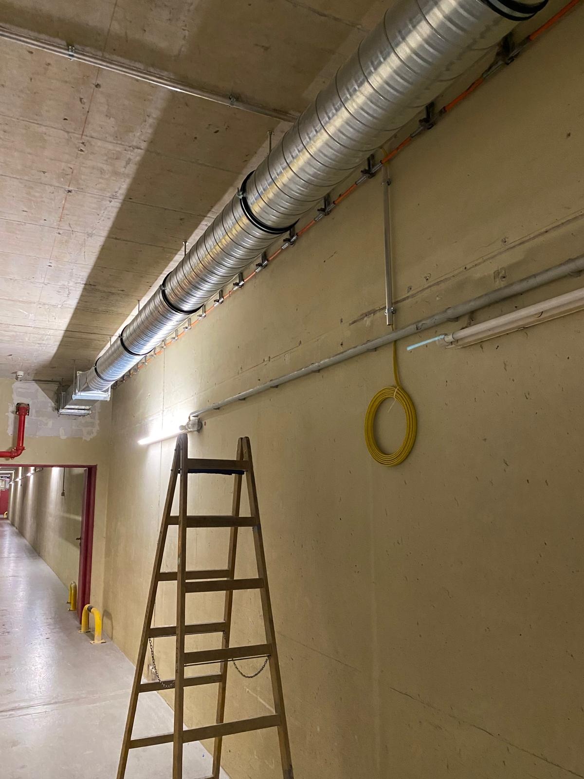 Montaż wentylacji w korytarzu budynku. Widoczna drabina, rury wentylacyjne pod sufitem, przewody elektryczne i oświetlenie. Surowy betonowy sufit i beżowe ściany.