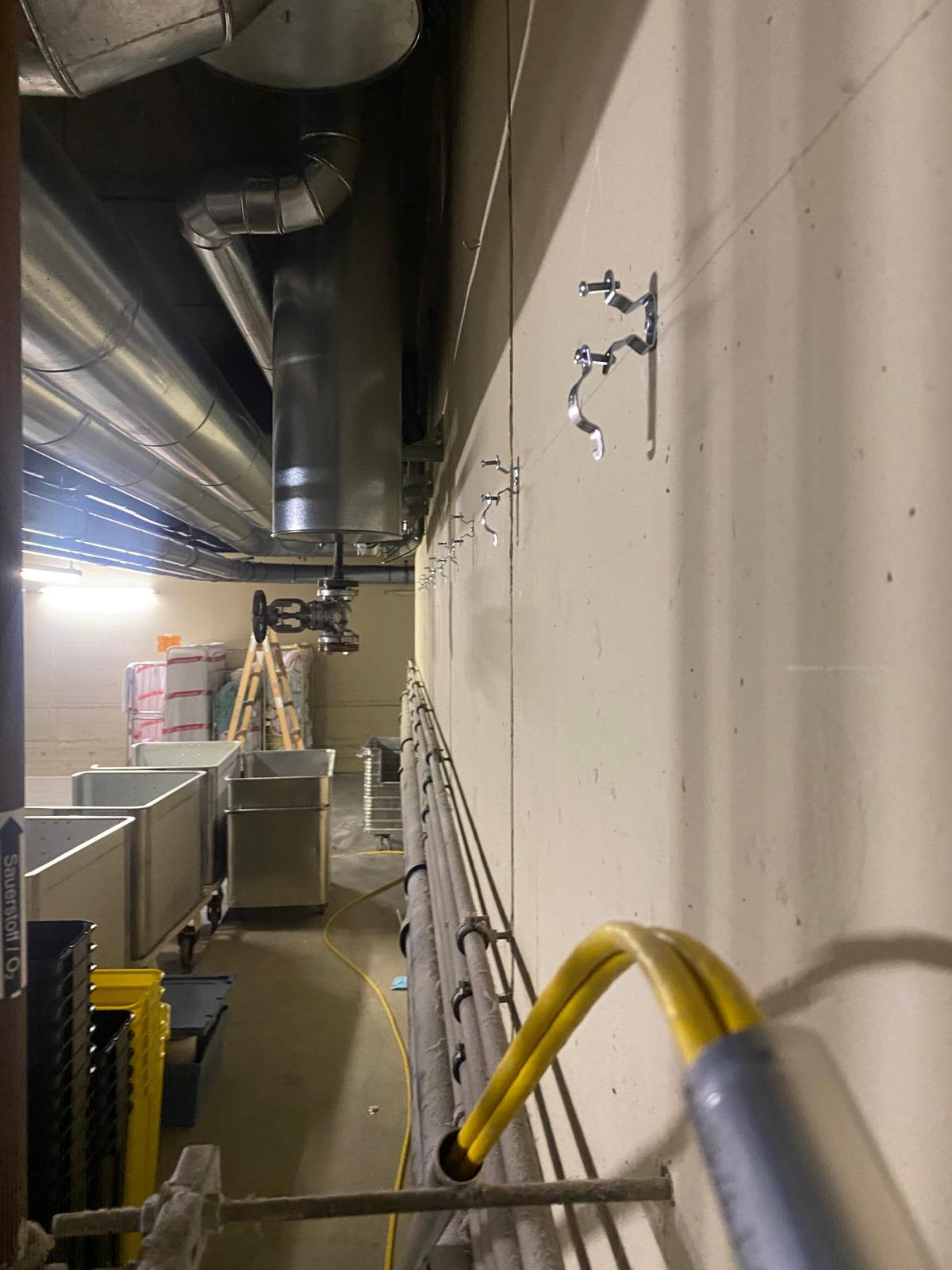 Rurociąg przemysłowy z zaworami i wspornikami na ścianie, widoczne kable w peszlu, drabina i wózki transportowe w tle. Ujęcie z dołu.