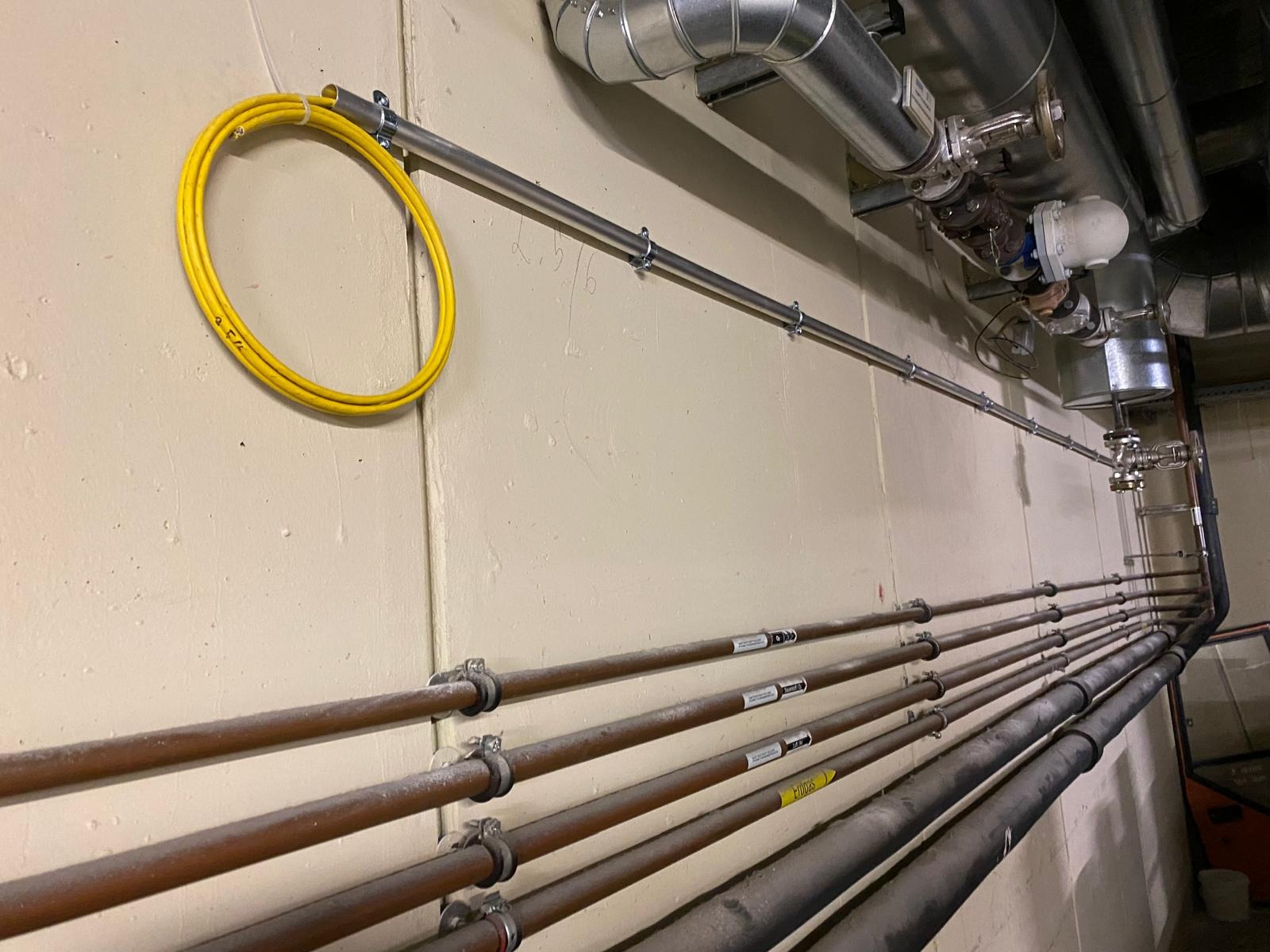 Rzędy rur miedzianych i stalowych, przymocowanych do ściany klamrami. U góry rura ocynkowana i zwój żółtego kabla. Widoczne etykiety na rurach.