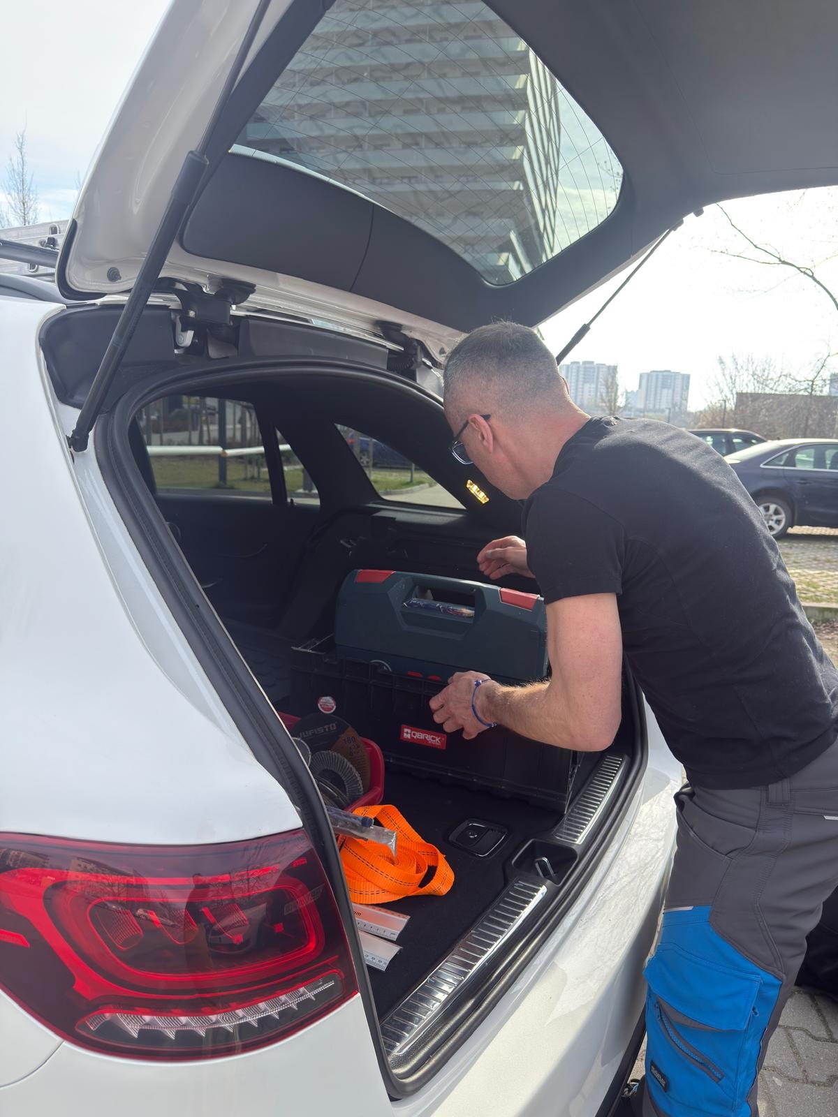Mężczyzna w czarnej koszulce sięga do bagażnika białego samochodu, gdzie widoczna skrzynka z narzędziami i akcesoriami. W tle budynek biurowy.