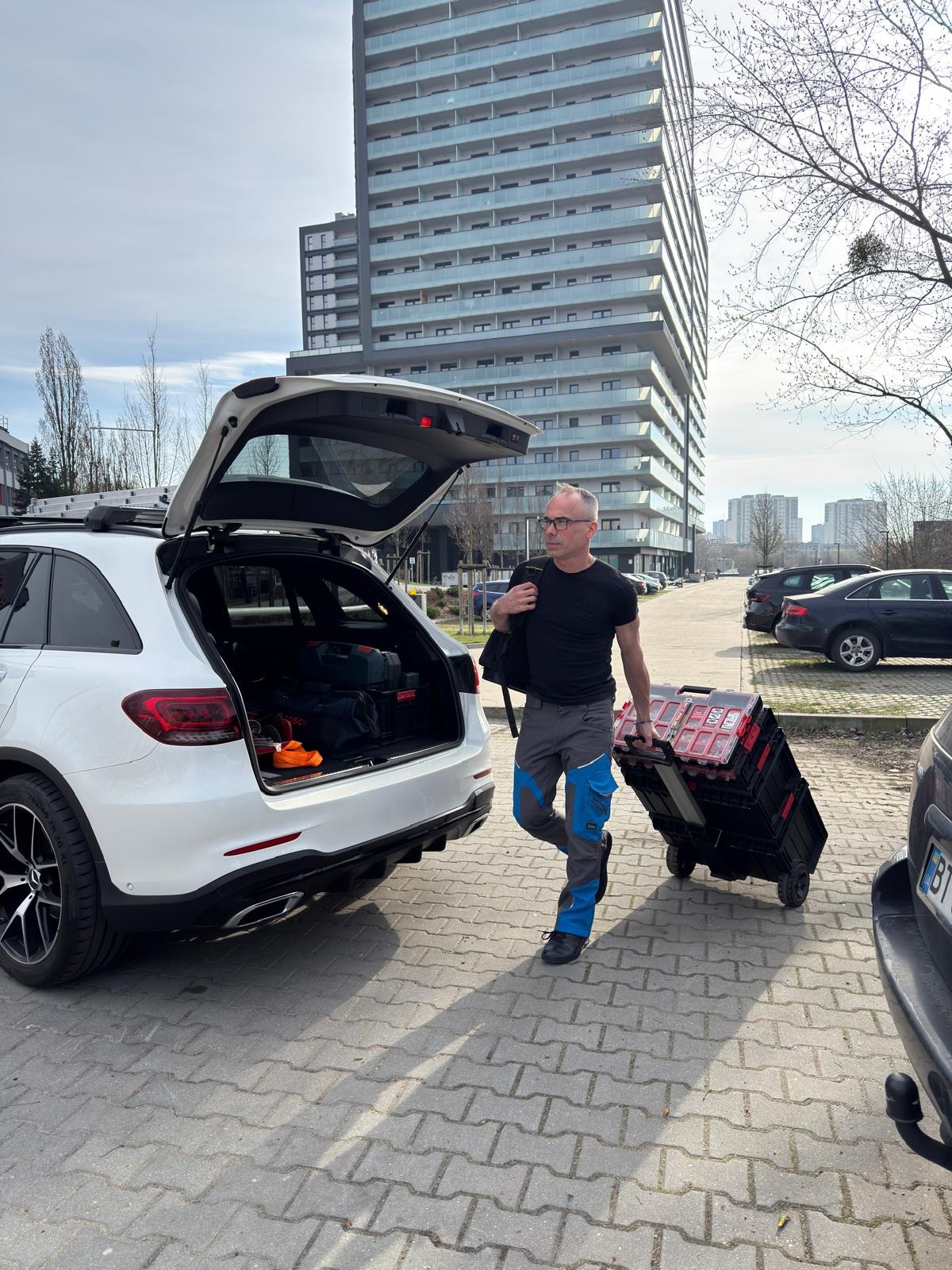 Fachowiec z walizką narzędzi idzie od samochodu z otwartym bagażnikiem, w tle budynek mieszkalny. Gotowość do pracy.